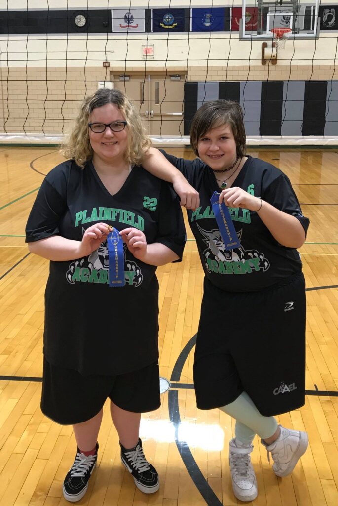 Two girls posing next to volleyball net holding award ribbons